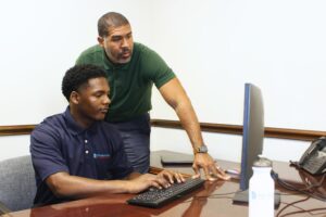 George Stewart stands beside intern Adam Tolliver, who is seated at a computer in the Foundation’s office, receiving guidance during a work session.
