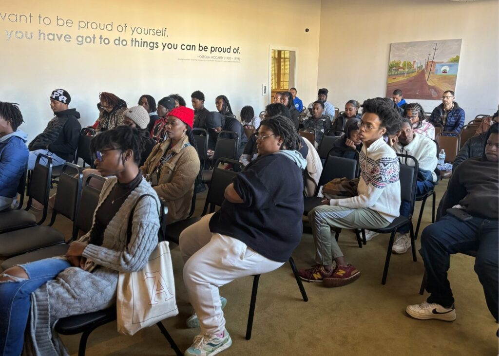 Community members of all ages seated in a meeting room, listening attentively to a speaker during a public gathering.