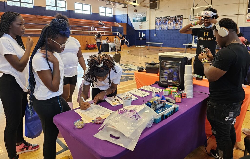 Large indoor career fair with rows of booths and tables set up in a gymnasium. Attendees, including students and job seekers, walk through the space, talk with recruiters, and review materials. Banners and displays from different organizations are visible in the background.