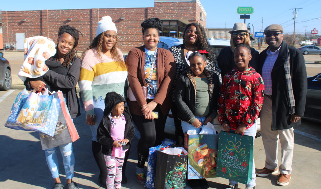 A group of adults and children stand outdoors in a parking lot, smiling and holding holiday gift bags. Some children wear festive outfits, and the group poses together on a sunny day.