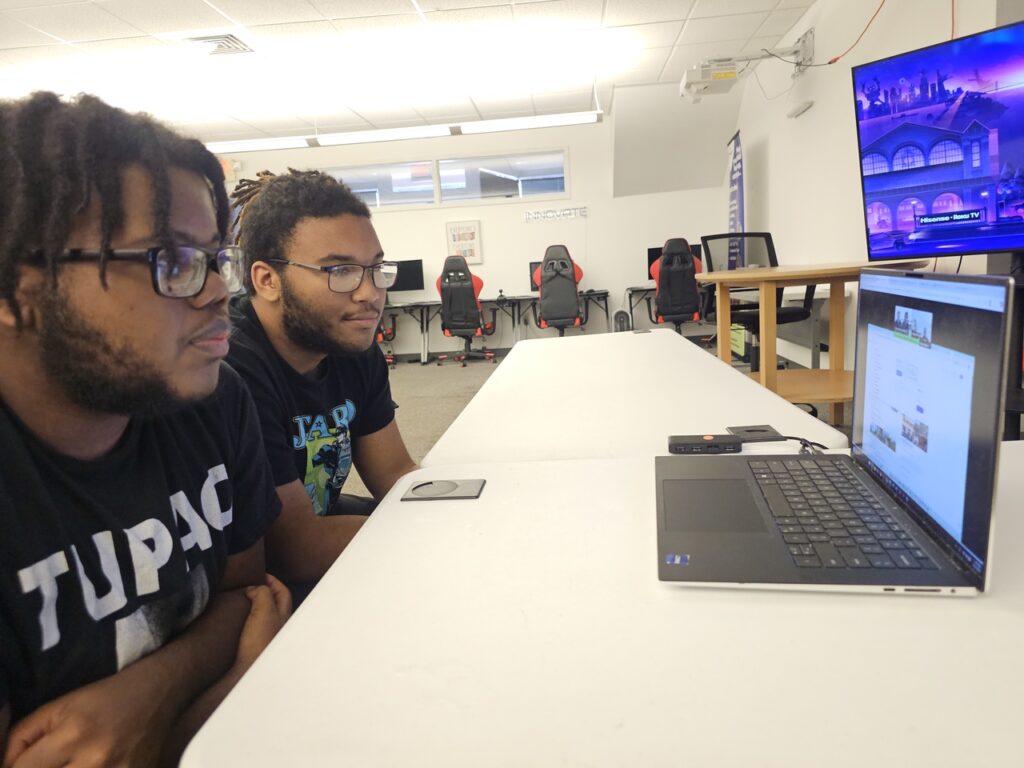 Two young men sit at a table, focused on a laptop screen during a virtual session. Behind them are gaming chairs and a large monitor displaying a colorful digital scene in a modern classroom or training space.
