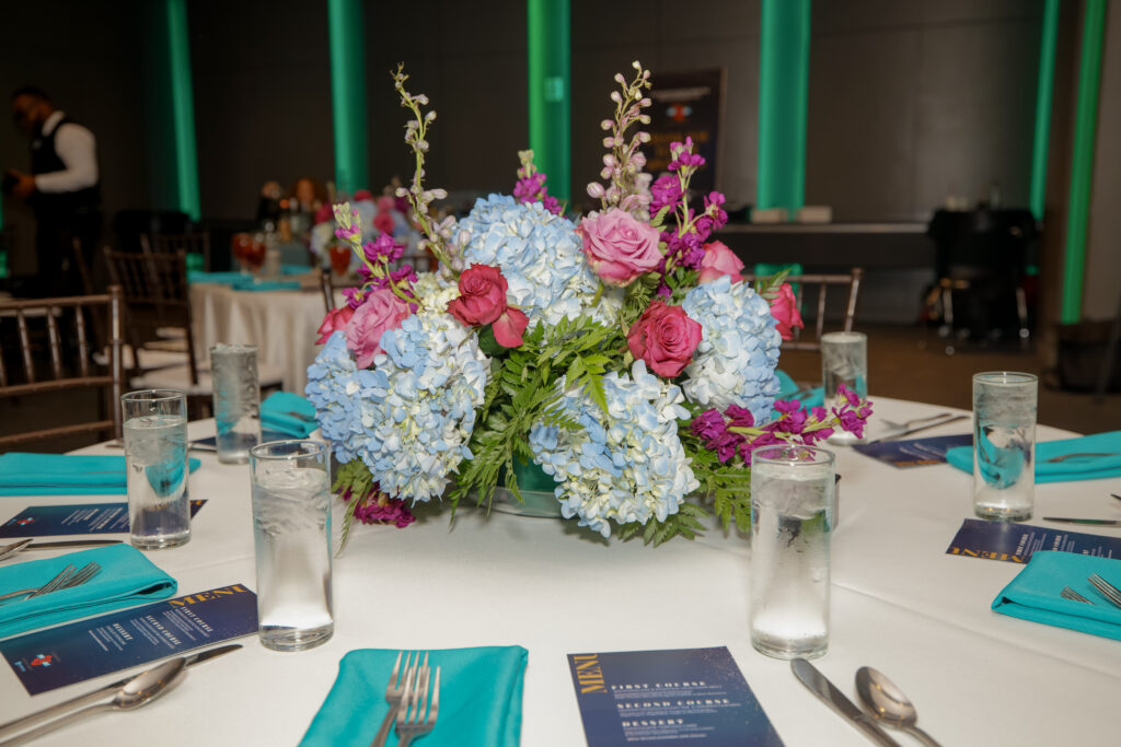 Floral centerpiece of blue hydrangeas, pink roses, and greenery arranged on a dinner table.