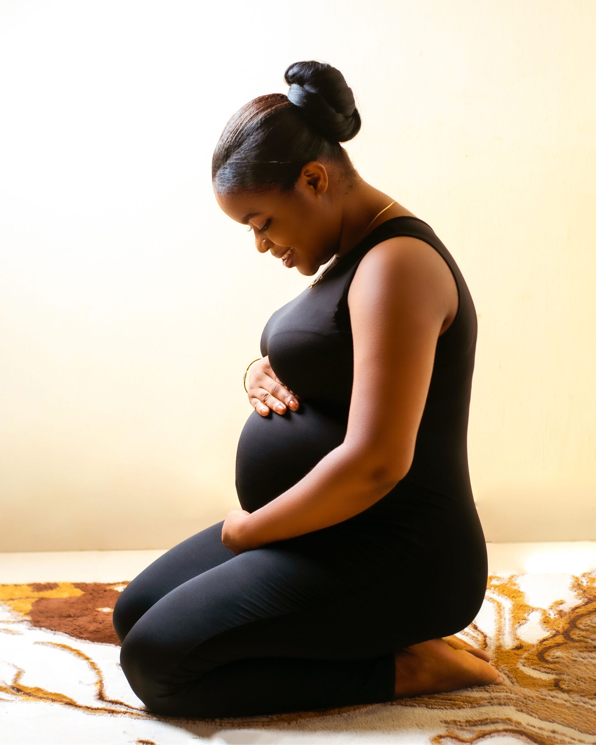 Pregnant Black woman kneeling indoors with her hands resting on her stomach.