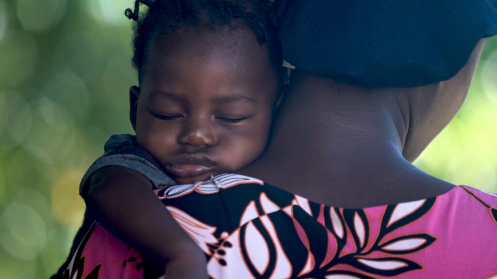 Young child sleeping on a mother's shoulder outdoors.